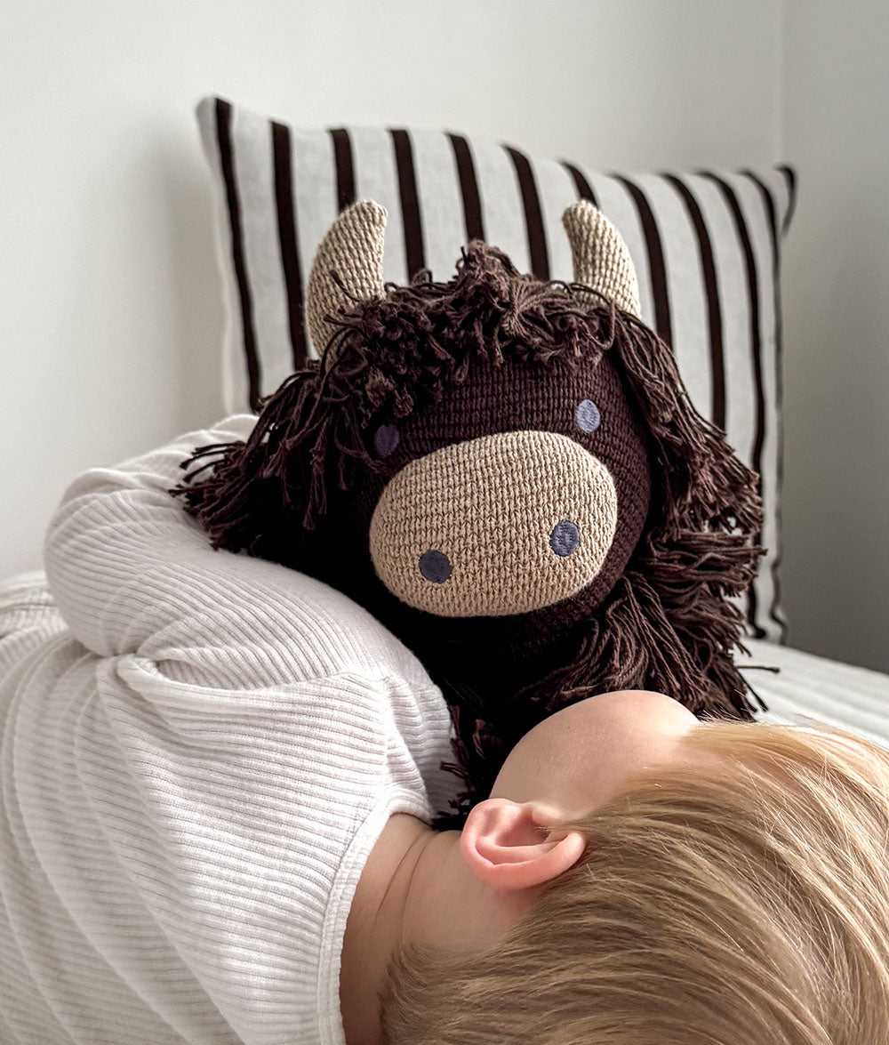 A close-up lifestyle shot of a child resting their head on a white ribbed blanket while hugging a dark brown and beige knitted cow plush toy. A black and white striped pillow is visible in the background.