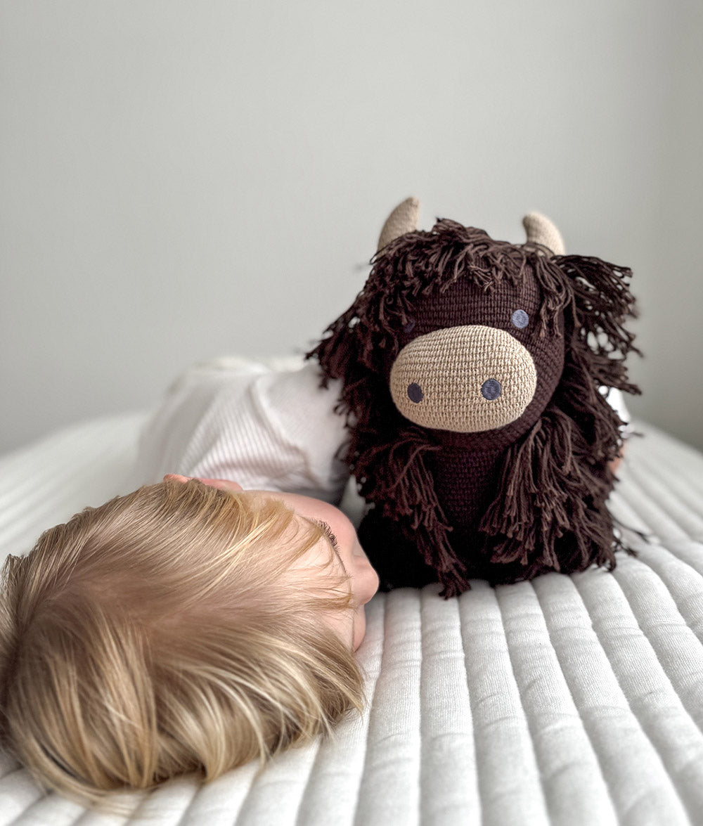 A close-up lifestyle shot of a child lying on a white quilted bed next to a dark brown shaggy crocheted highland cow toy with a beige snout and small horns.