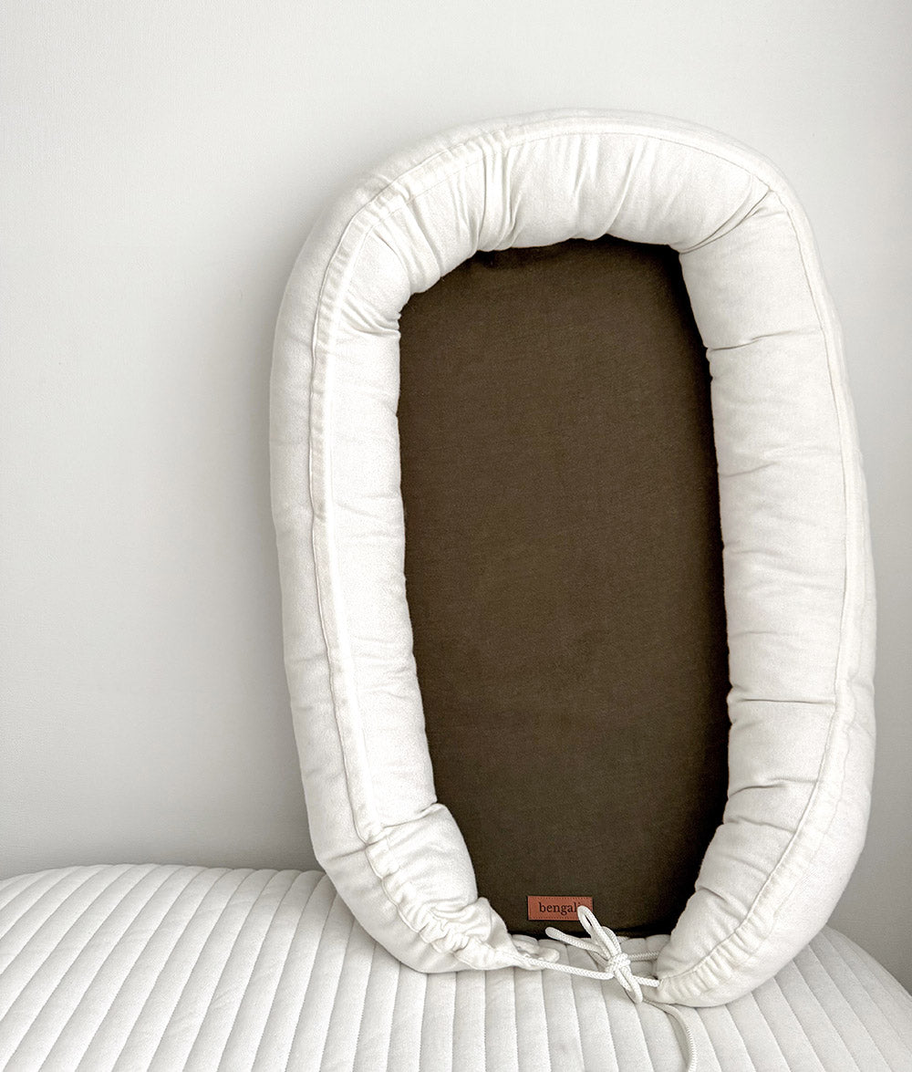 A padded baby lounger with a white cushioned border and a olive green center panel, resting on a white ribbed blanket against a light gray wall.