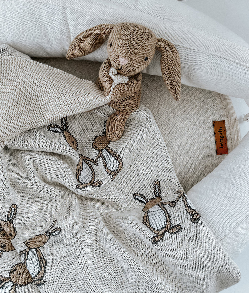 A beige and brown bunny-shaped comforter snuggled in a white blanket with a pillow, placed on a neutral background.