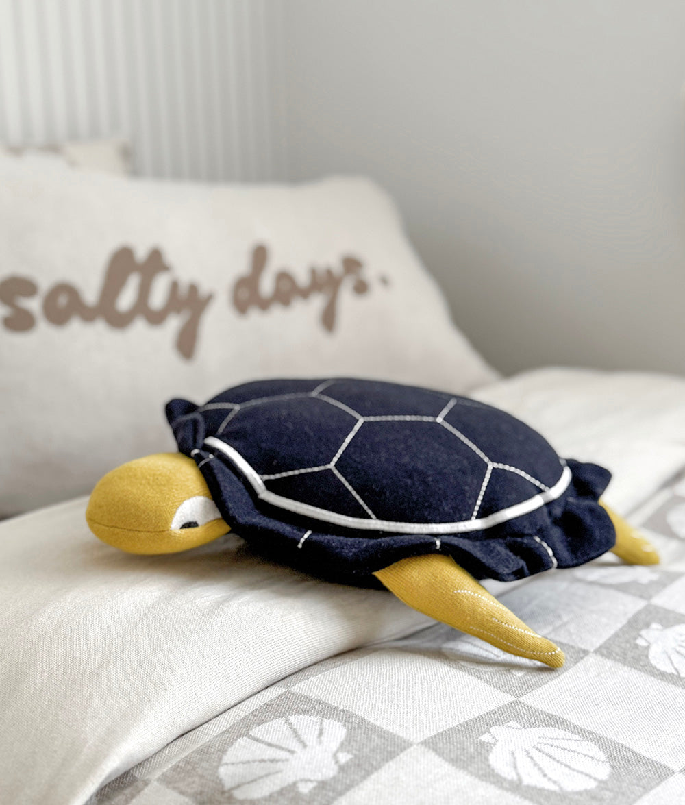 Turtle-shaped pillow on a bed with 'salty days' pillow in the background