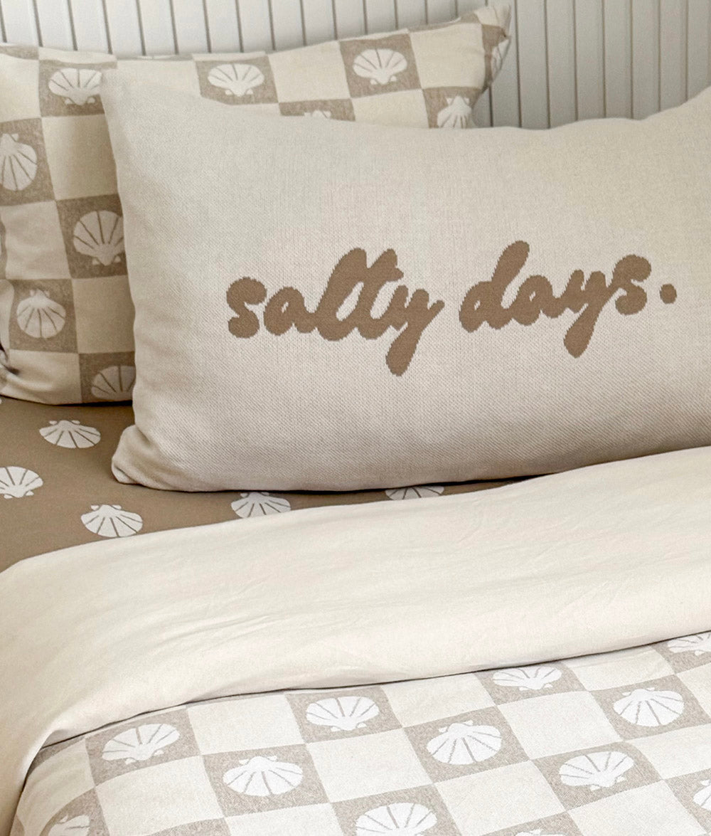 Beige pillow with 'salty days' text on a bed with shell-patterned bedding.
