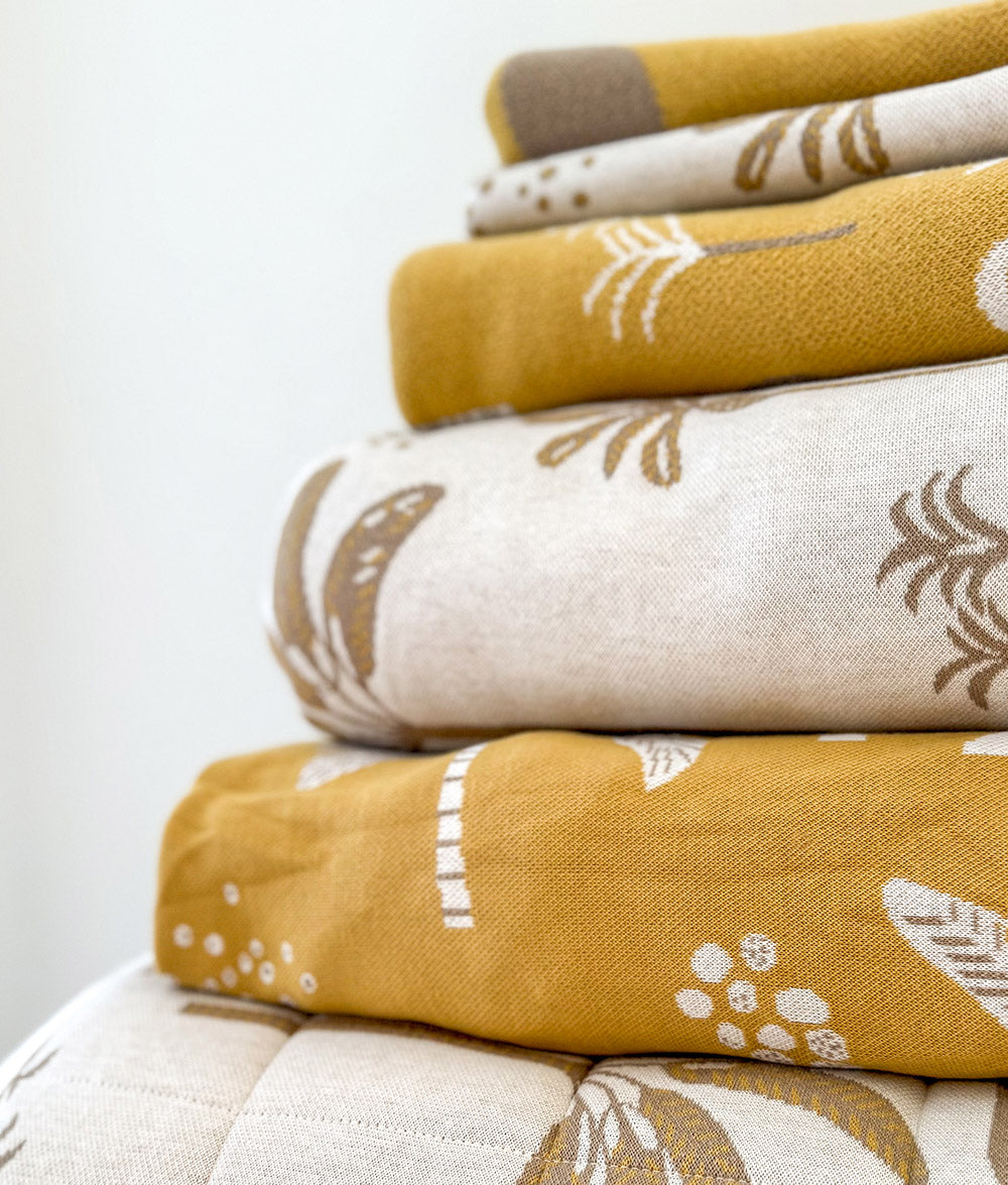 A stack of folded cotton textiles, blankets and bedding, in mustard yellow and ivory with brown and white palm tree and botanical leaf patterns.