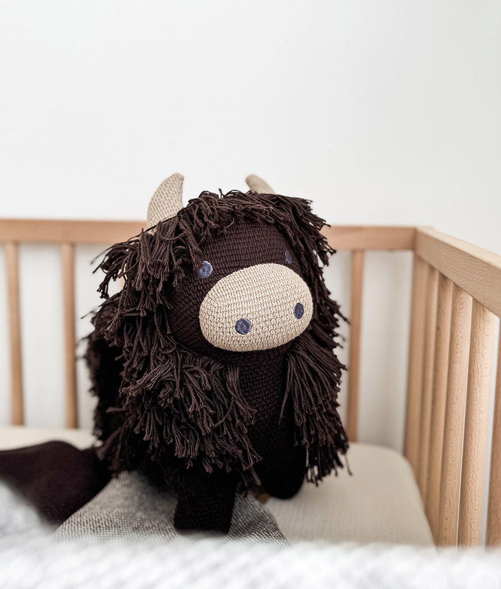 A dark brown crocheted highland toy plush toy with shaggy dark brown hair, a cream-colored snout, and small cream horns, sitting inside a light wood crib.