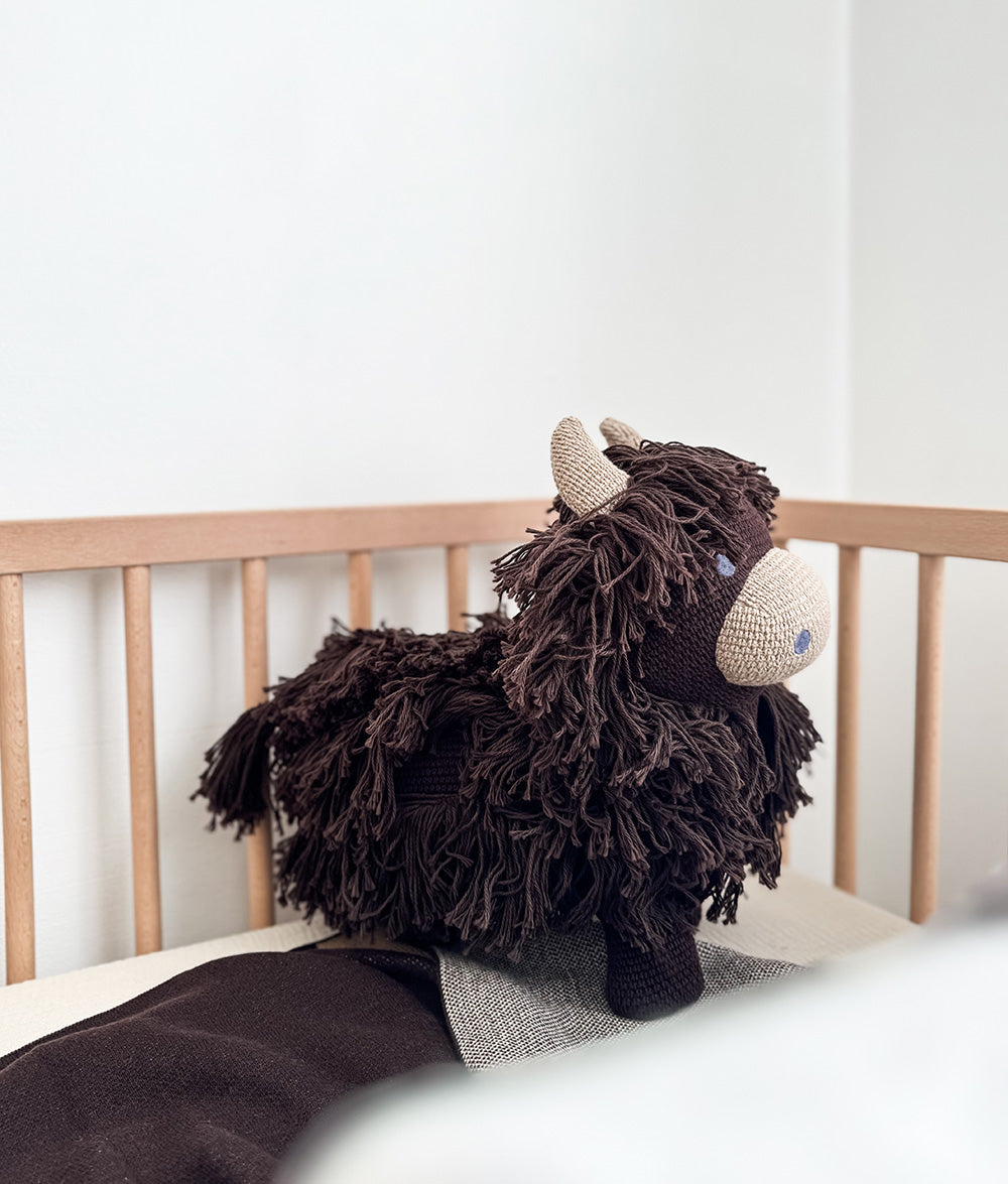 A dark brown shaggy knit stuffed highland toy with cream horns and a cream snout, sitting on a wooden bed against a white wall.