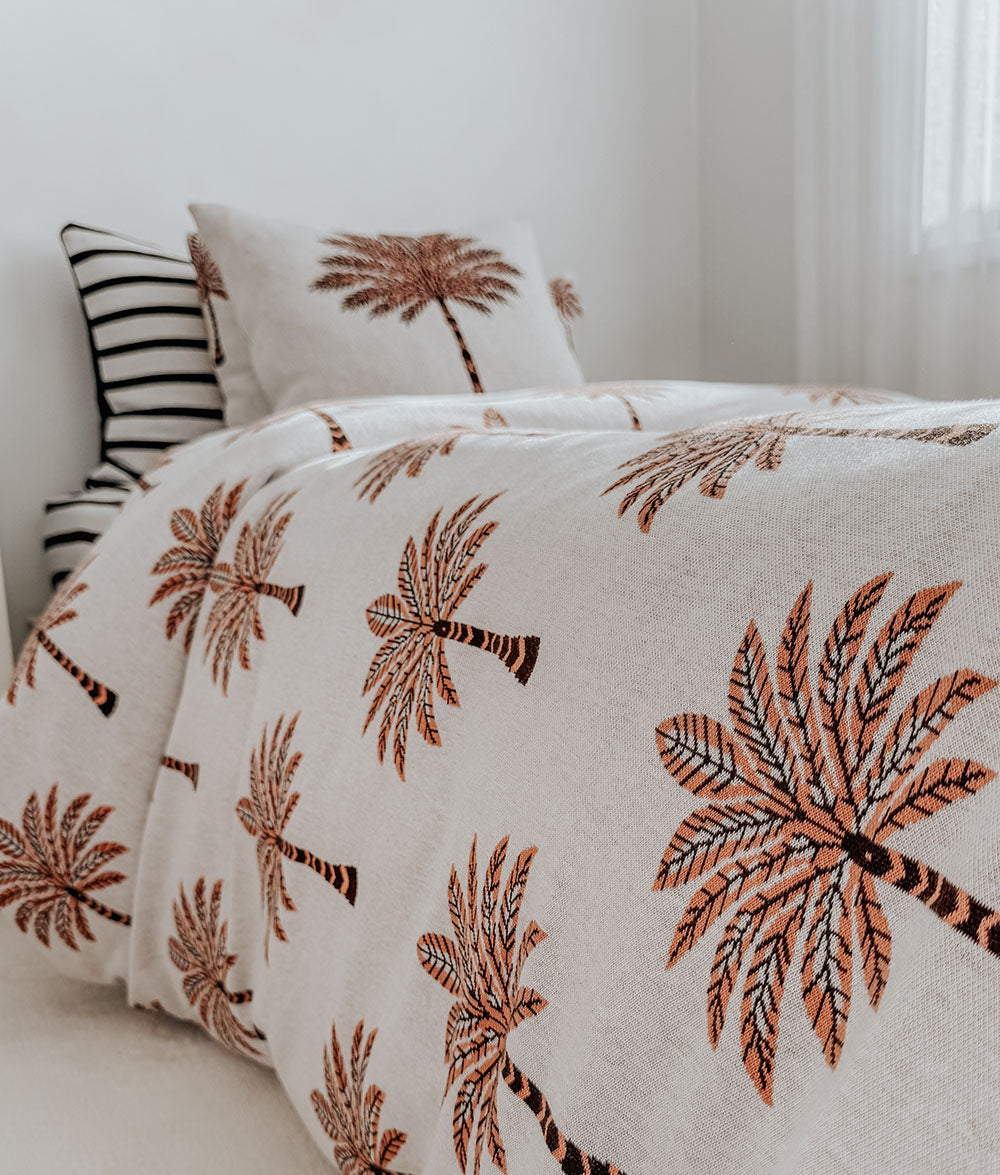 A close-up of a bed dressed in a cream-colored duvet cover and pillowcase featuring a repeating embroidered or printed palm tree pattern in terracotta and dark brown, with a black-and-white striped accent pillow behind it.