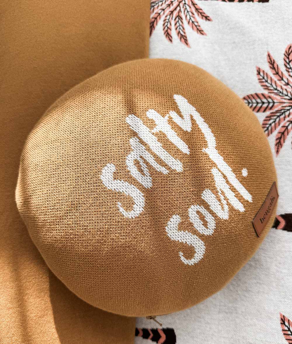A round tobacco knit filled cushion with white text reading 'Salty soul.' and a small brown brand tag, shown on matching Peach Sundown bedding.