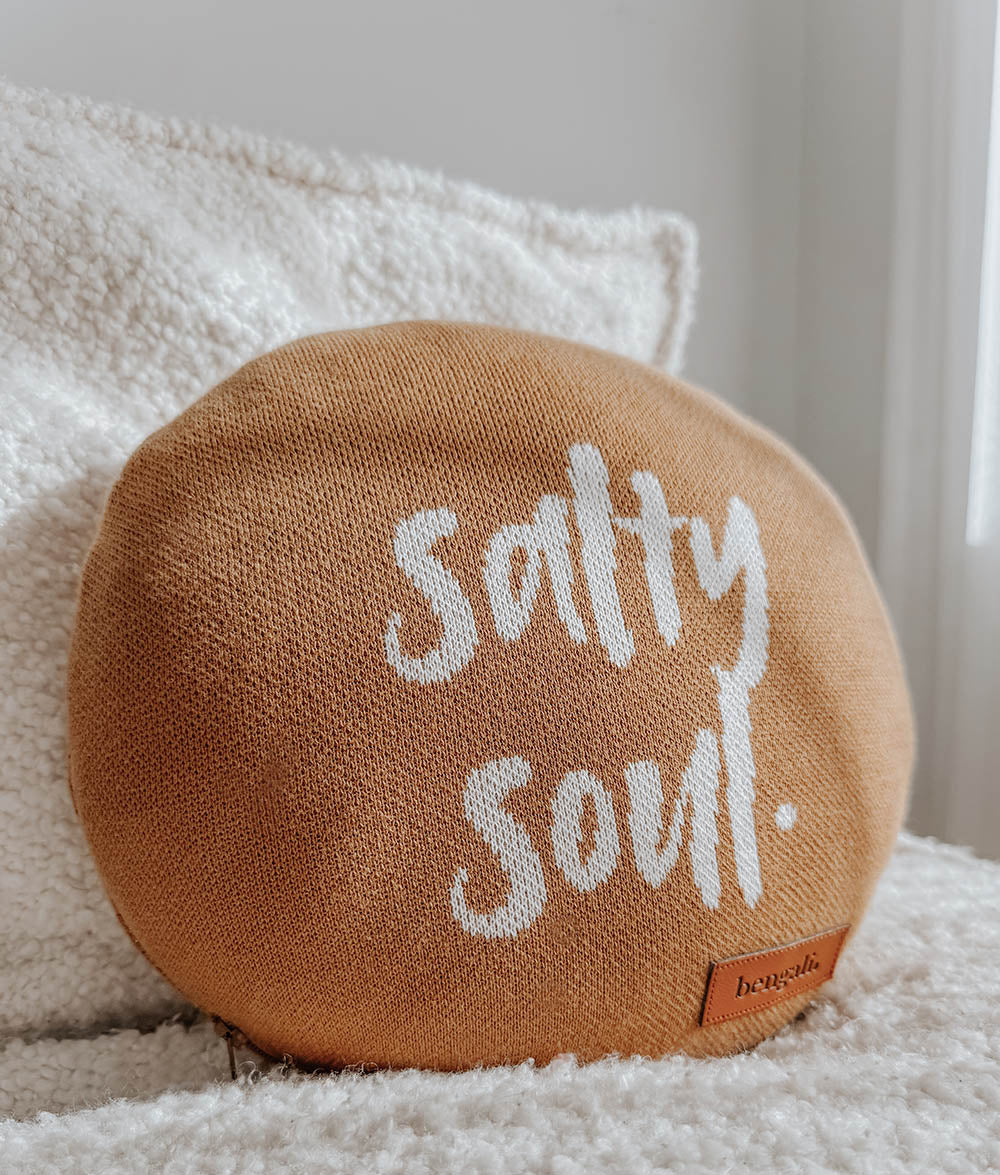 A round tobacco tan throw pillow with the words 'Salty Soul.' knitted in white across the front, resting on a white textured chair.
