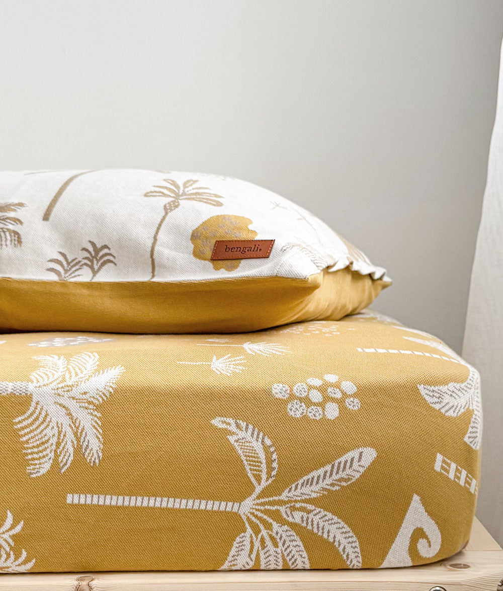 A close-up of a yellow and white patterned cushion or pillow set, with a smaller matching pillow resting on top of a single bed. The fabric has a tropical palm leaf and wave print, and a small brown 'bengali' brand tag is visible on the top pillow.