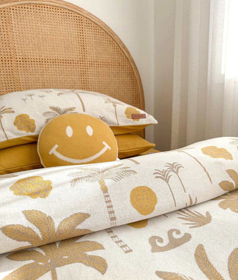 A close-up of a bed styled with a ivory-colored tropical print bedding set featuring palm trees and yellow wave motifs, a matching yellow smiley face pillow, and a woven cane headboard.