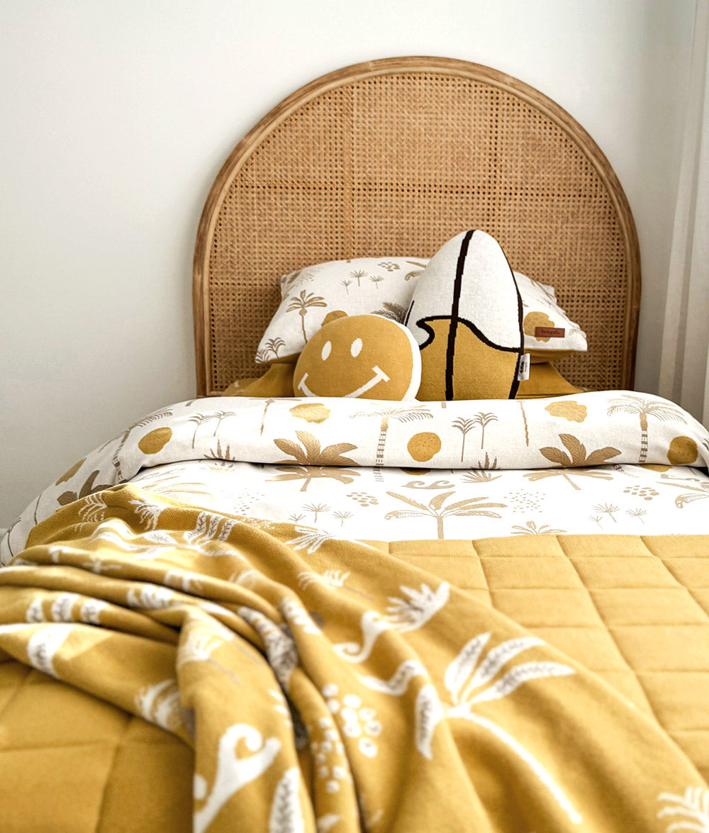 A cozy bed styled with a mustard yellow quilted blanket, a matching patterned duvet cover, a woven cane headboard, and decorative pillows and pillowcases.