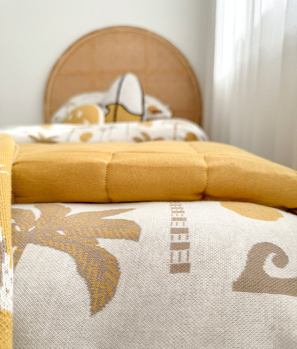 A close-up of a bed styled with a mustard yellow quilted blanket and an ivory duvet cover featuring a palm tree and wave pattern, with a round woven headboard in the background.