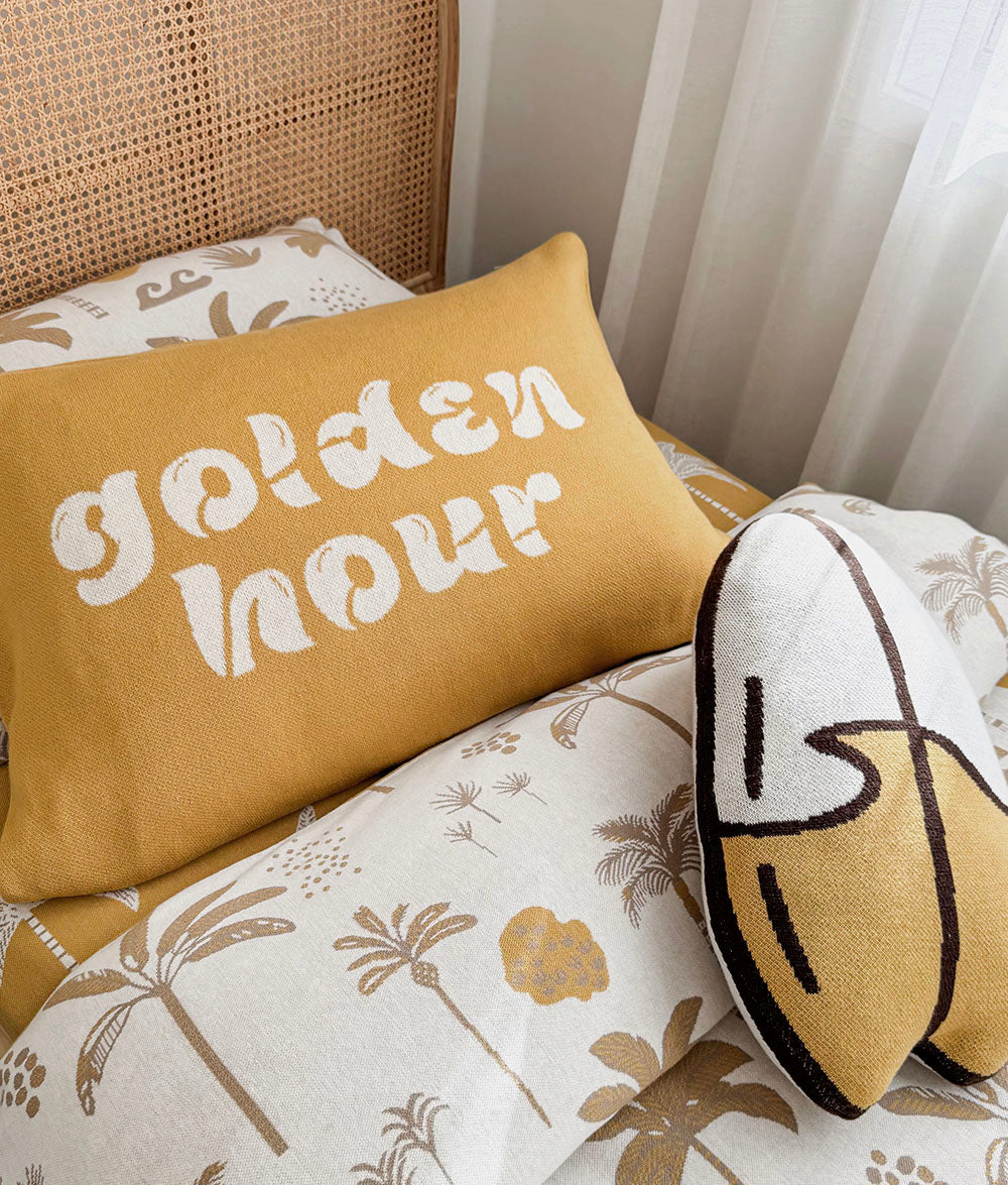 A styled bedroom scene with a mustard yellow pillow featuring the words 'golden hour' in white script, layered on top of a cream and brown palm tree patterned duvet. A second abstract yellow surfboard cushion with chocolate lines rests on the right, and a woven cane headboard and white curtain are visible in the background.