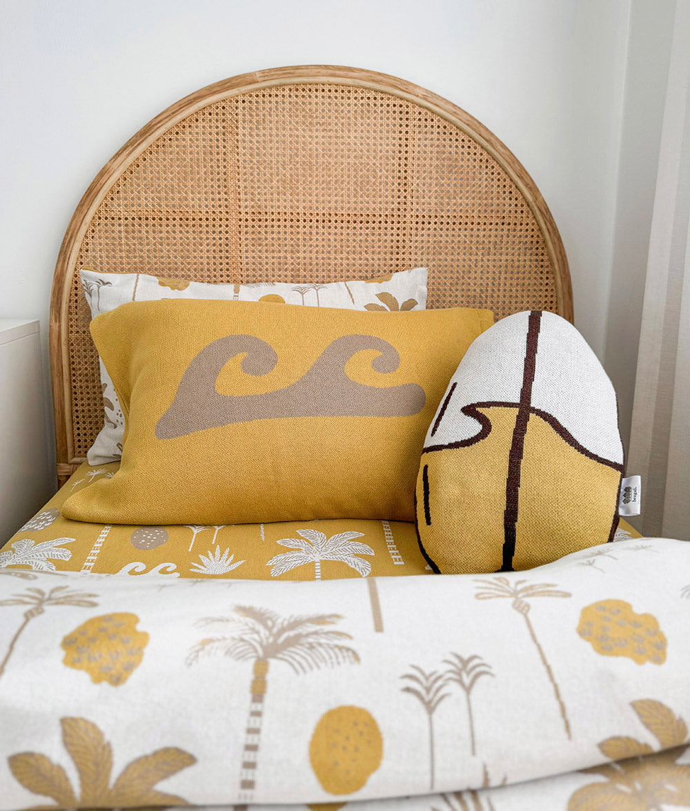 A styled bed featuring a mustard yellow pillow with a gray wave design, a white pillow with a palm print, and a large round surfboard decorative pillow, set against a natural woven cane headboard and a white tropical-patterned duvet.