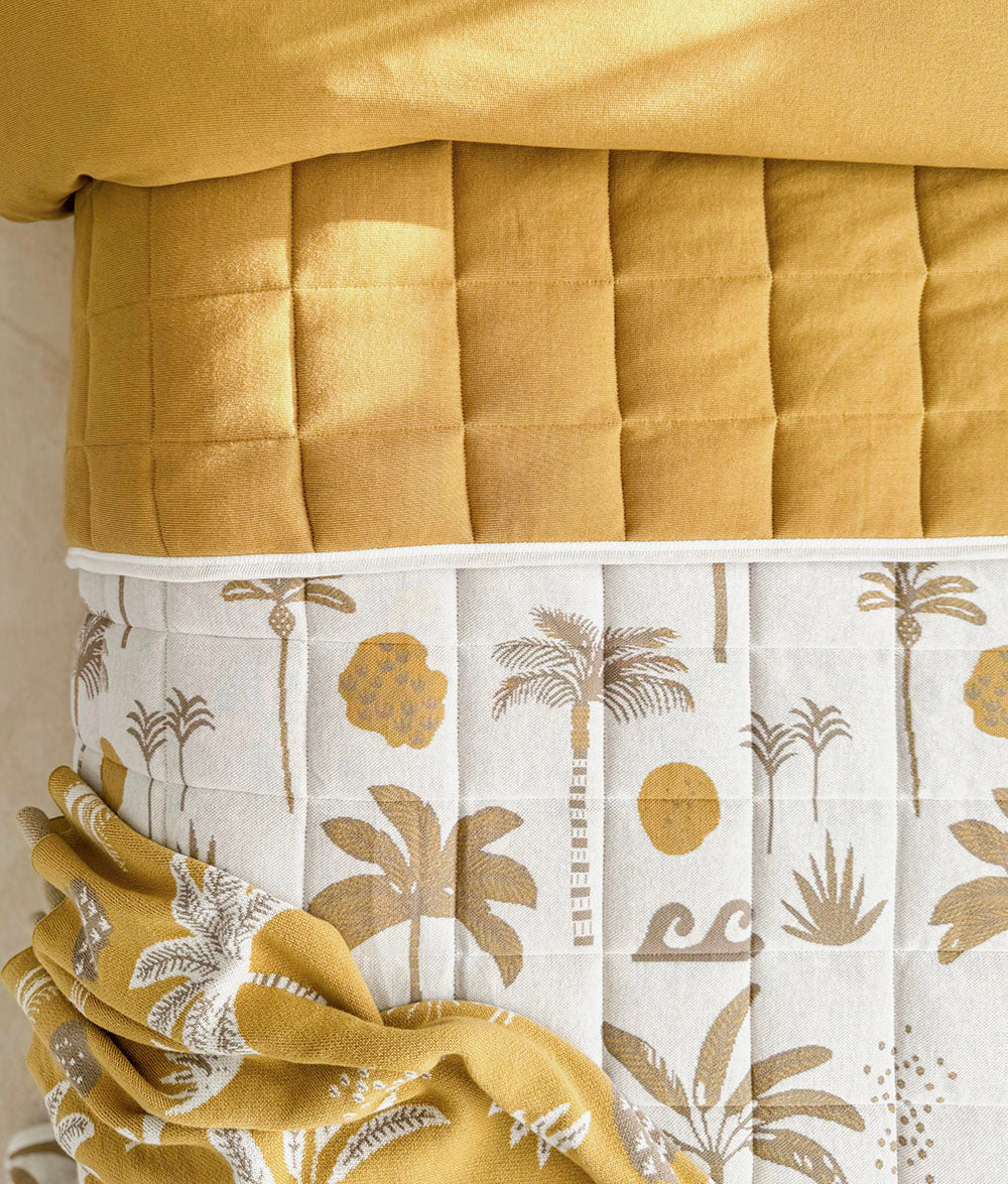A close-up of a quilted bedding set in a warm mustard yellow and white tropical print. The top section features a solid mustard yellow quilted duvet folded over, while the lower section displays a white quilted panel with a gold and gray palm tree and botanical pattern, accented by a matching mustard yellow golden hour blanket gathered in the lower left corner.