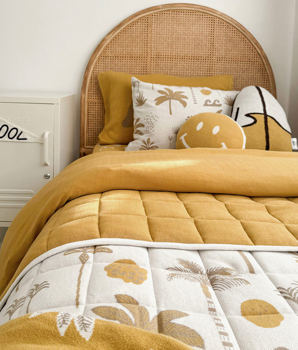 A cozy bed styled with a mustard yellow quilted blanket, matching pillows, and a white quilt featuring a tan palm tree pattern, set against a light wood cane headboard.