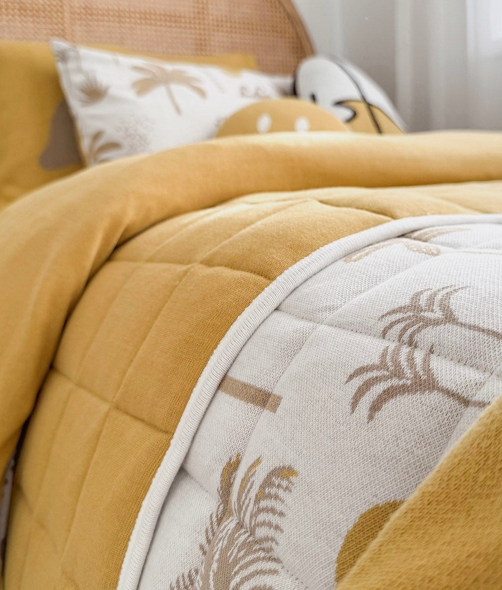 A close-up of a quilted bedding set in mustard yellow and off-white, featuring a palm tree pattern on the throw and matching pillows.