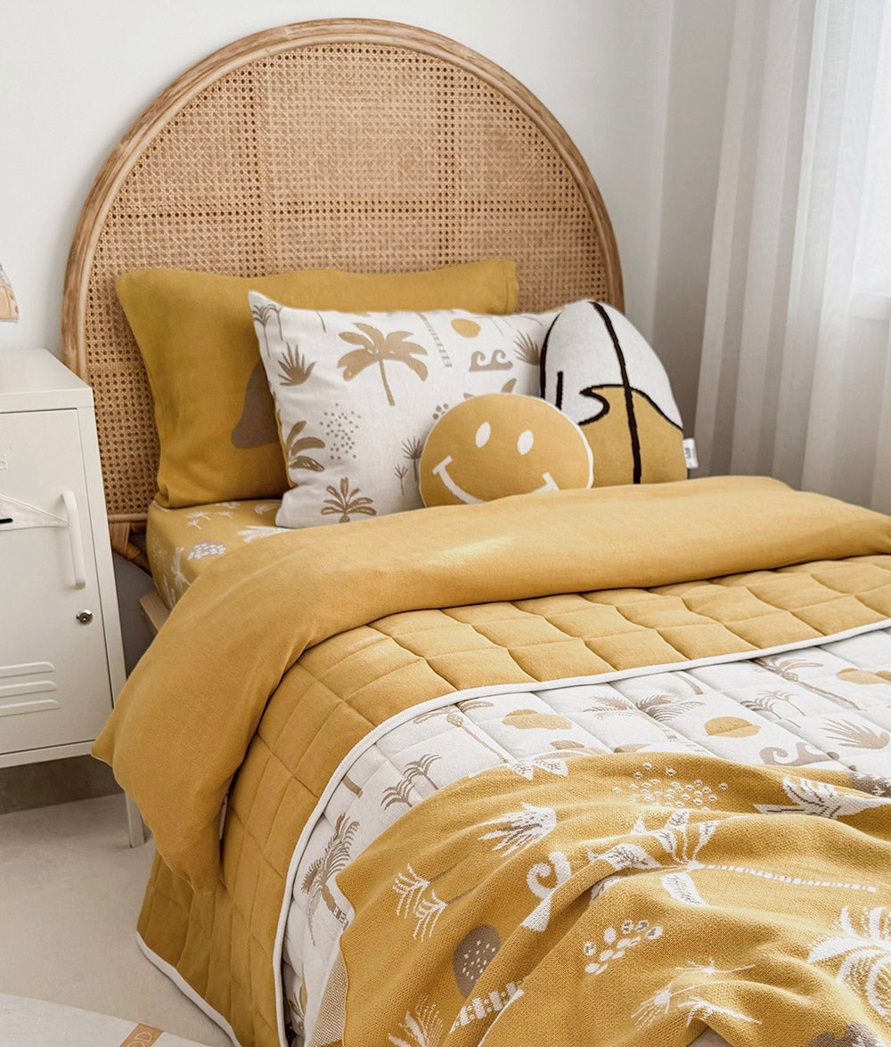 A cozy bed styled with a mustard yellow quilted comforter, matching pillow, patterned white and beige pillows, and a woven cane headboard in a bright bedroom setting.