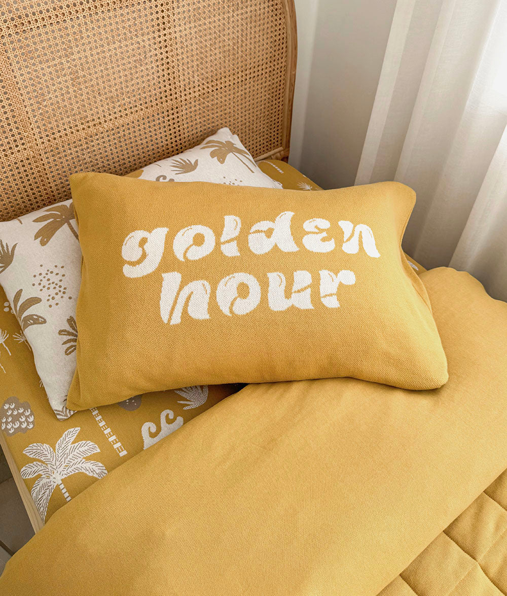 A mustard yellow rectangular throw pillow with the words 'golden hour' in white script, styled on a bed with a matching yellow quilt and a patterned cream pillow against a woven cane headboard.
