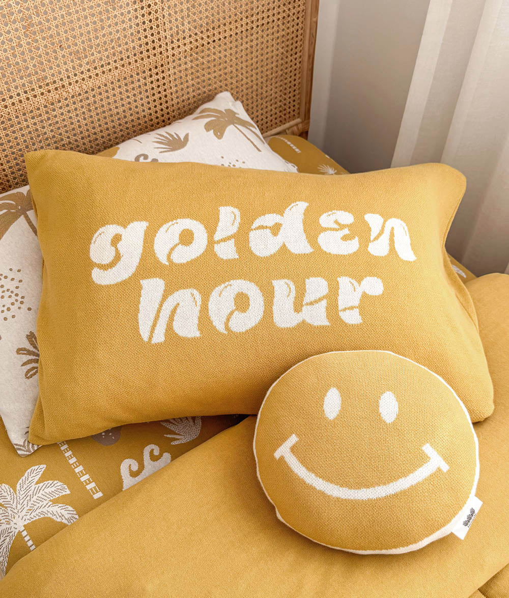 A yellow throw pillow with the words 'golden hour' in white script, styled on a bed with a matching round smiley face pillow and patterned linens.