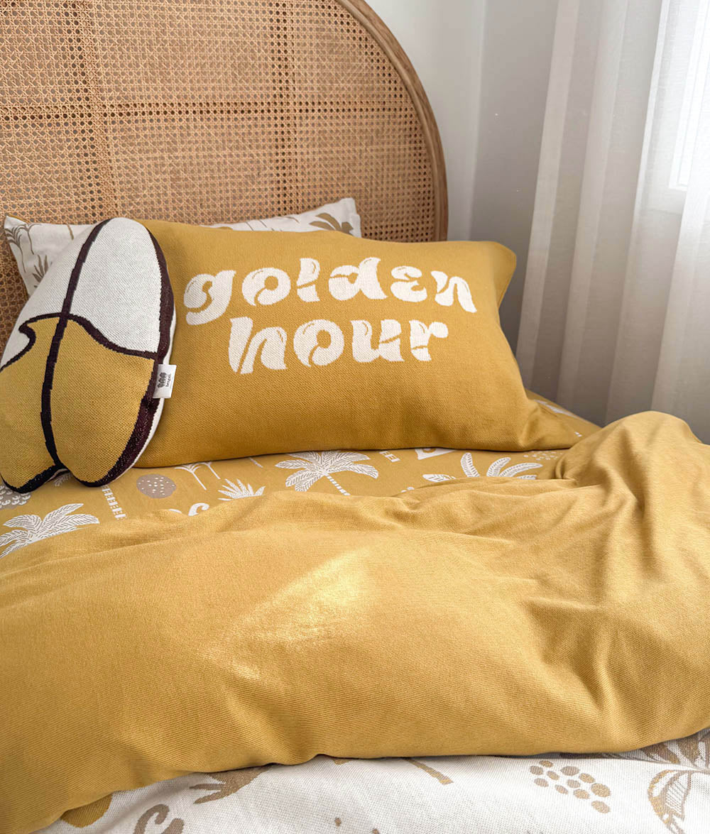 A cozy bedroom scene with a mustard yellow duvet and matching pillow featuring the words 'golden hour' in white script, alongside a patterned accent pillow and a woven cane headboard.
