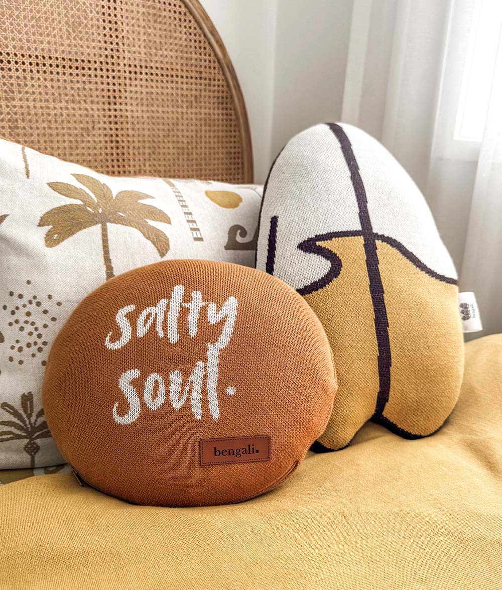 Two decorative throw pillows on a yellow bedspread in a bedroom setting. The foreground pillow is round, tobacco, and features the words 'salty soul.