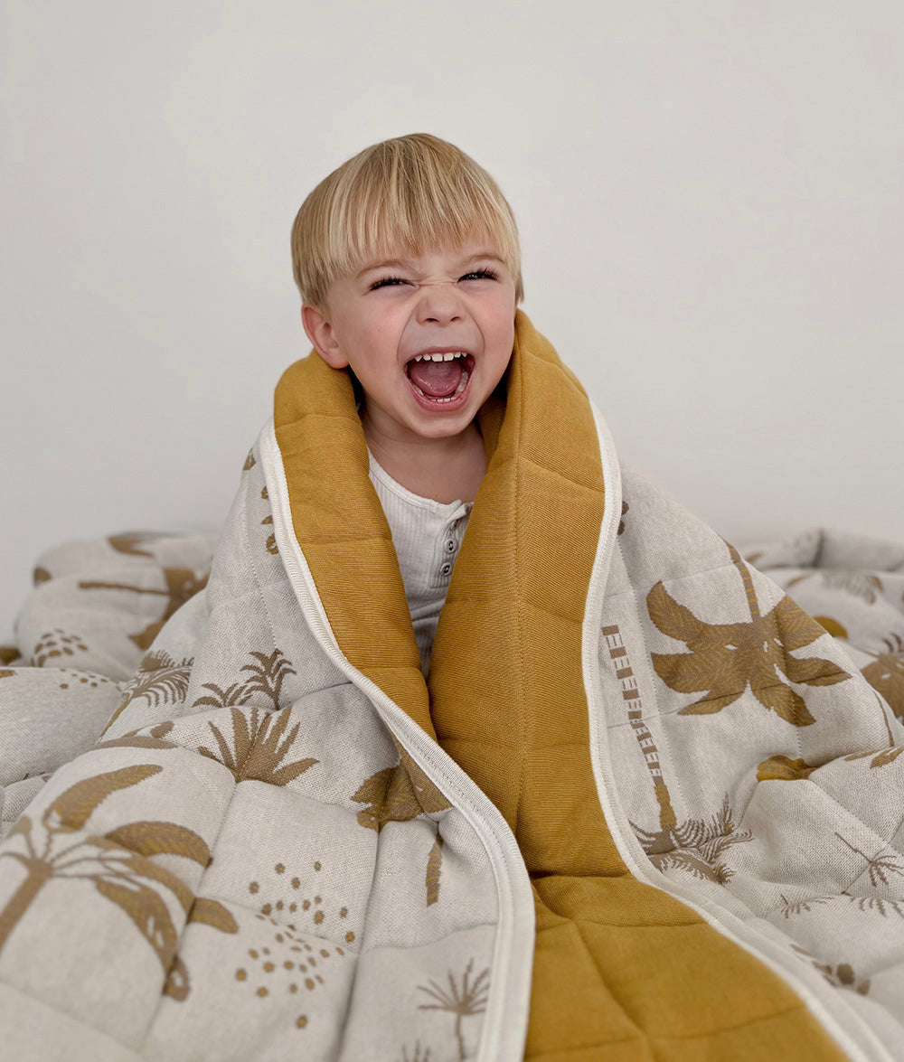 Child wrapped in the Golden Hour reversible quilt featuring an ivory base with golden mustard and beige coastal palm and wave motifs, with a plain golden mustard reverse.