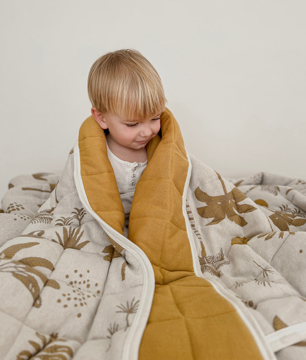 Child sitting wrapped in the Golden Hour reversible quilt with ivory coastal print and golden mustard reverse.