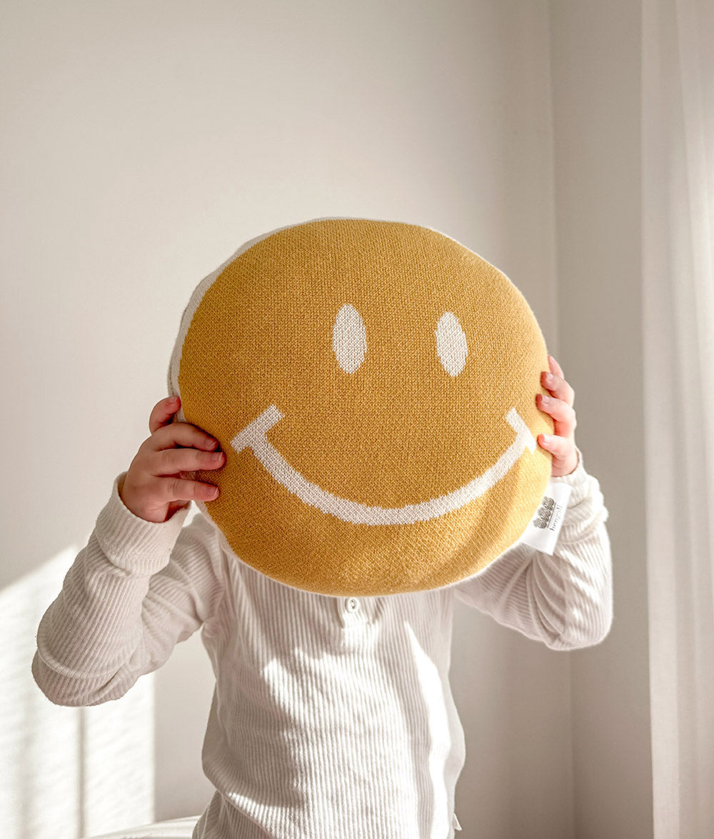 A child in a white ribbed long-sleeve shirt holds a round yellow knit smiley face pillow in front of their face against a softly lit cream wall.