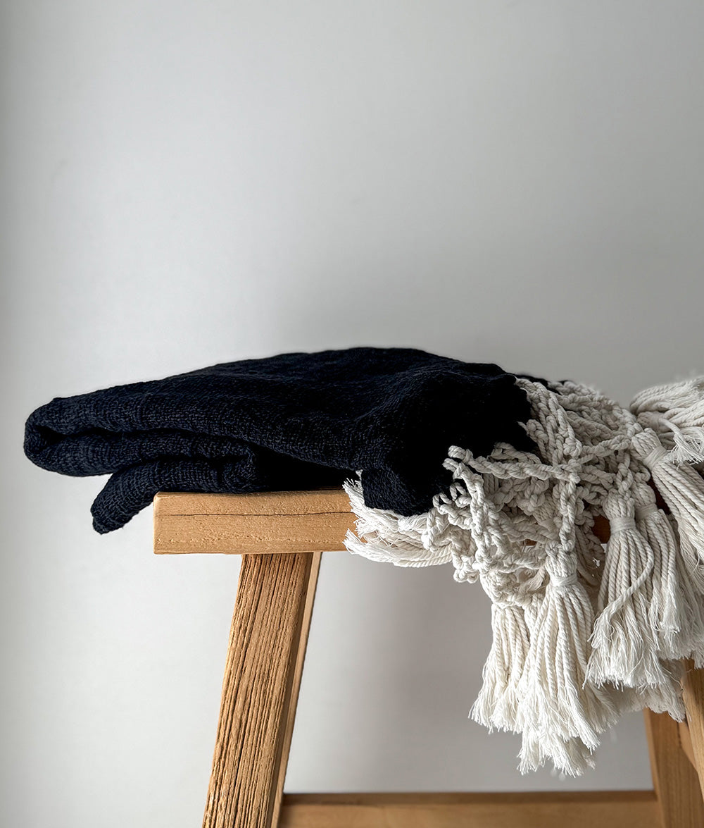 Black folded blanket and white textured throw on a wooden stool against a plain background