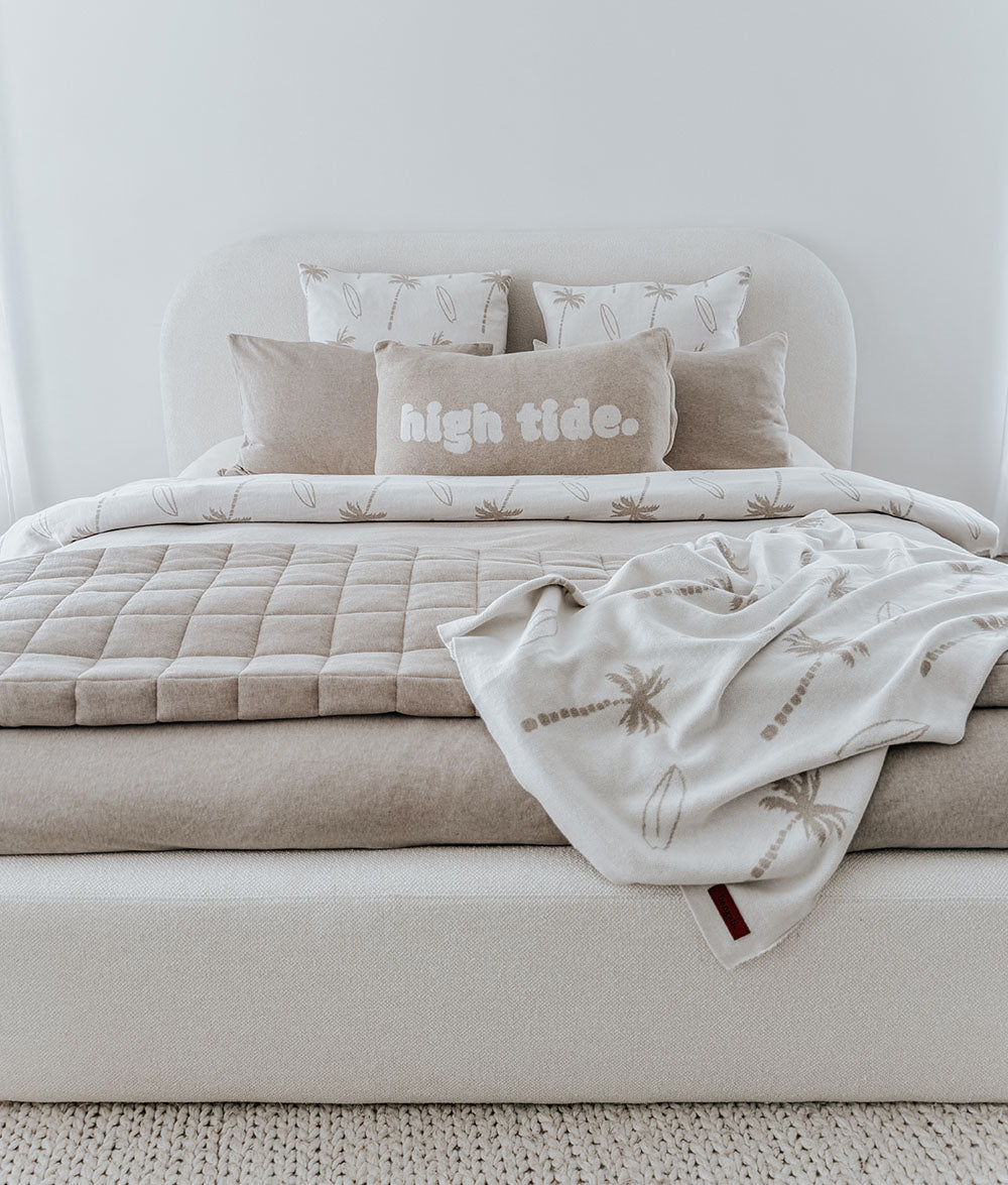 Neatly made bed with beige bedding and 'high tide.' pillow, featuring a towel draped over the foot of the bed.