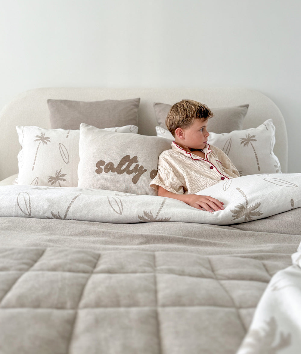 Child lying on a bed with 'salty days' pillow, surrounded by neutral-toned bedding and pillows.