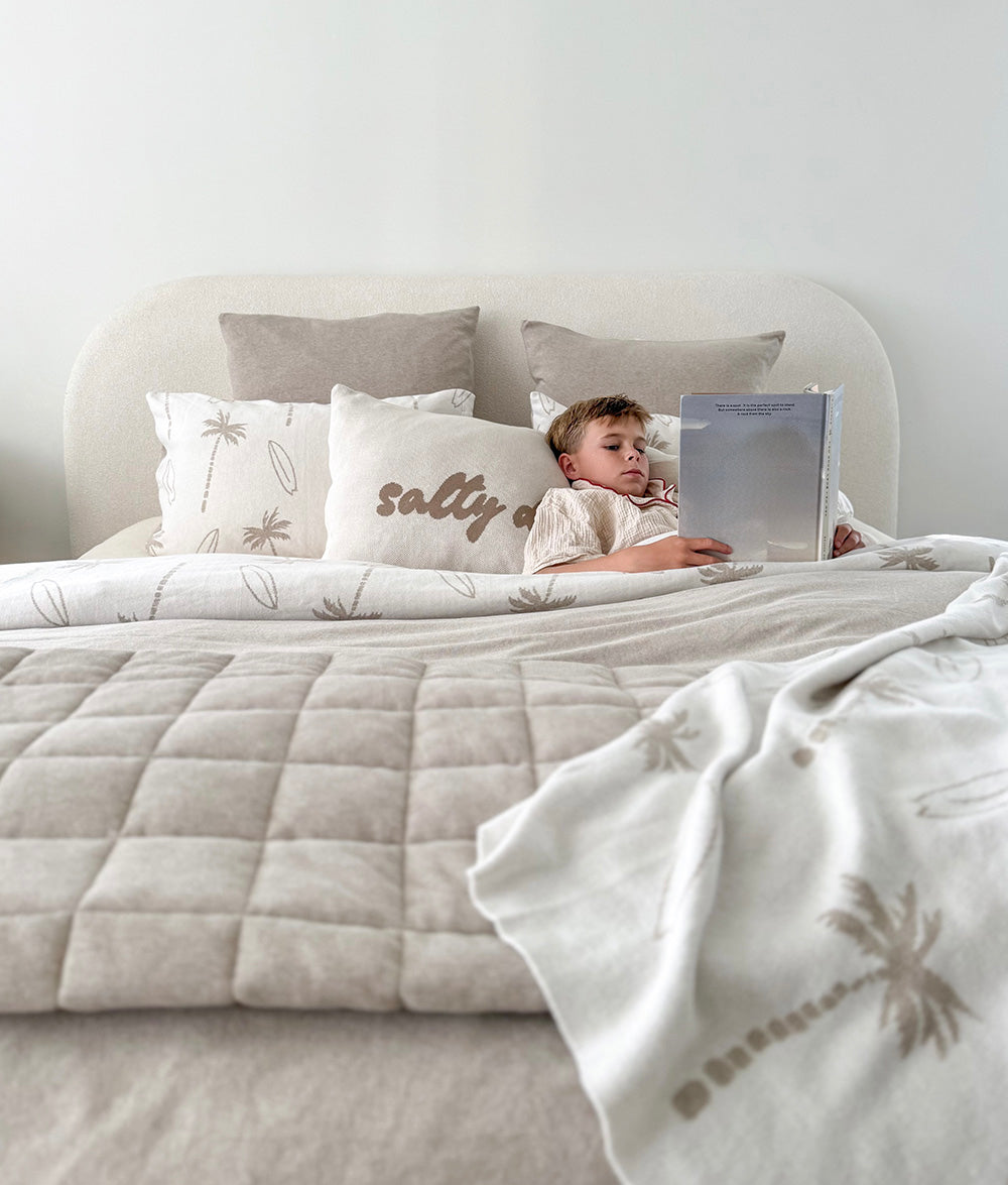 Child lying on a bed with a blanket and pillows, reading a book.