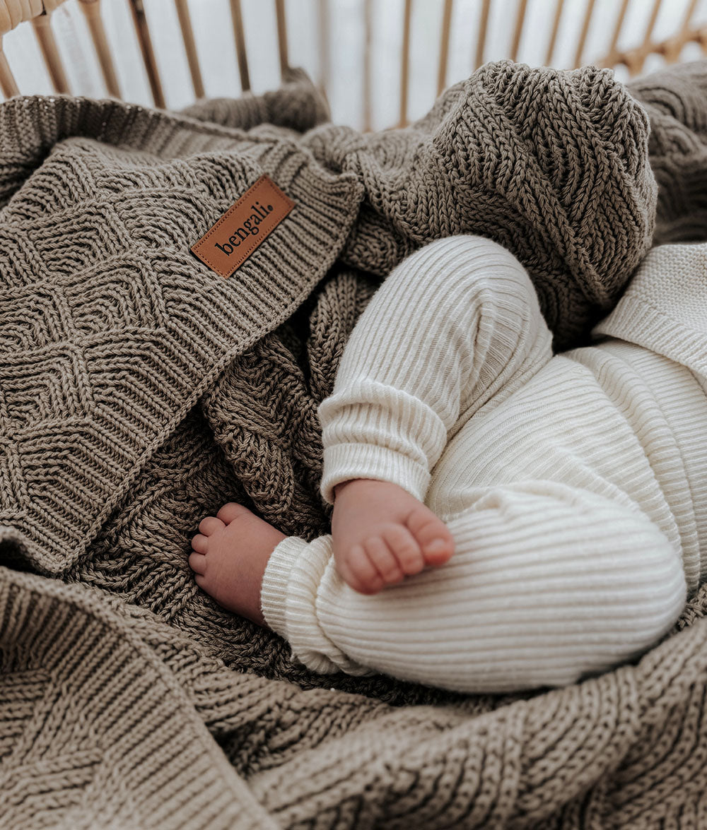 Baby wrapped in a white knitted outfit under a textured brown blanket with a brand label.