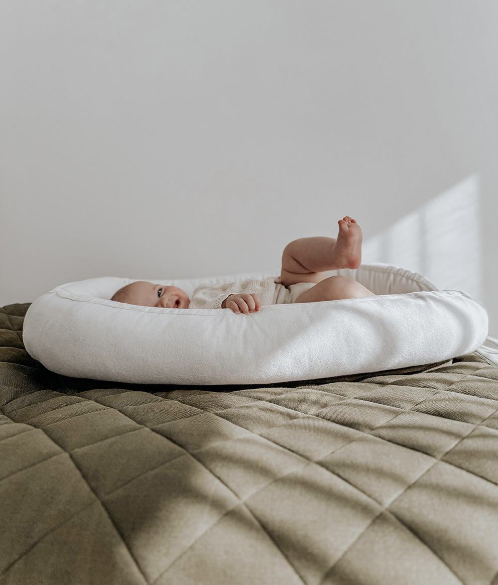 A baby lying on their back inside a white crescent-shaped bassinet or bed placed on top of a quilted olive green blanket, with a plain light gray wall in the background.