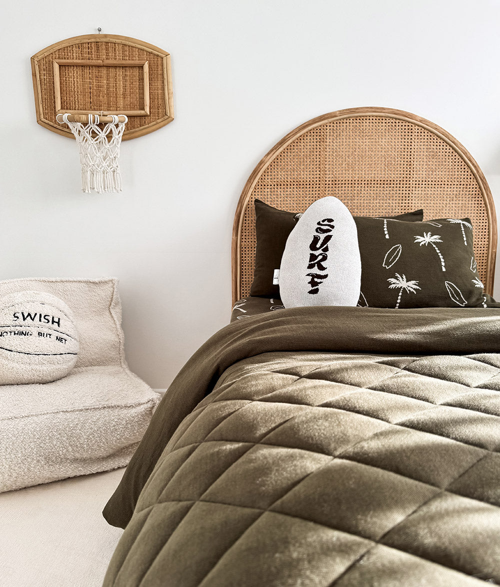 A styled bedroom scene with a bed featuring a natural woven rattan headboard, olive green quilted bedding, dark brown and white decorative pillows, and a small wall-mounted basketball hoop. A matching textured pouf with a pillow reading 'SWISH' is visible on the left.
