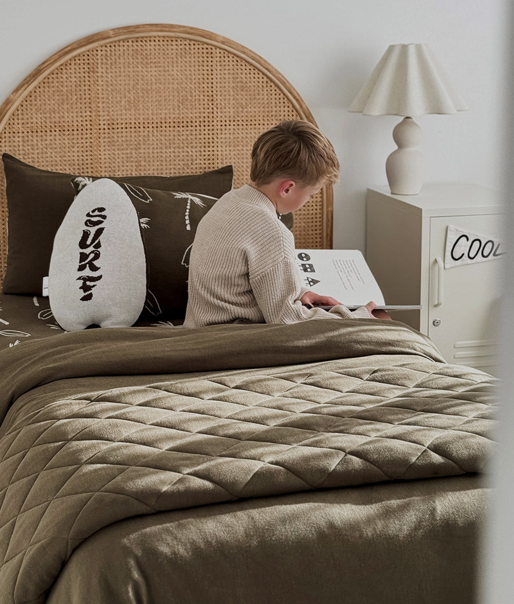 Child reading on a bed with Olive Surfing Palm bedding and matching cushions.