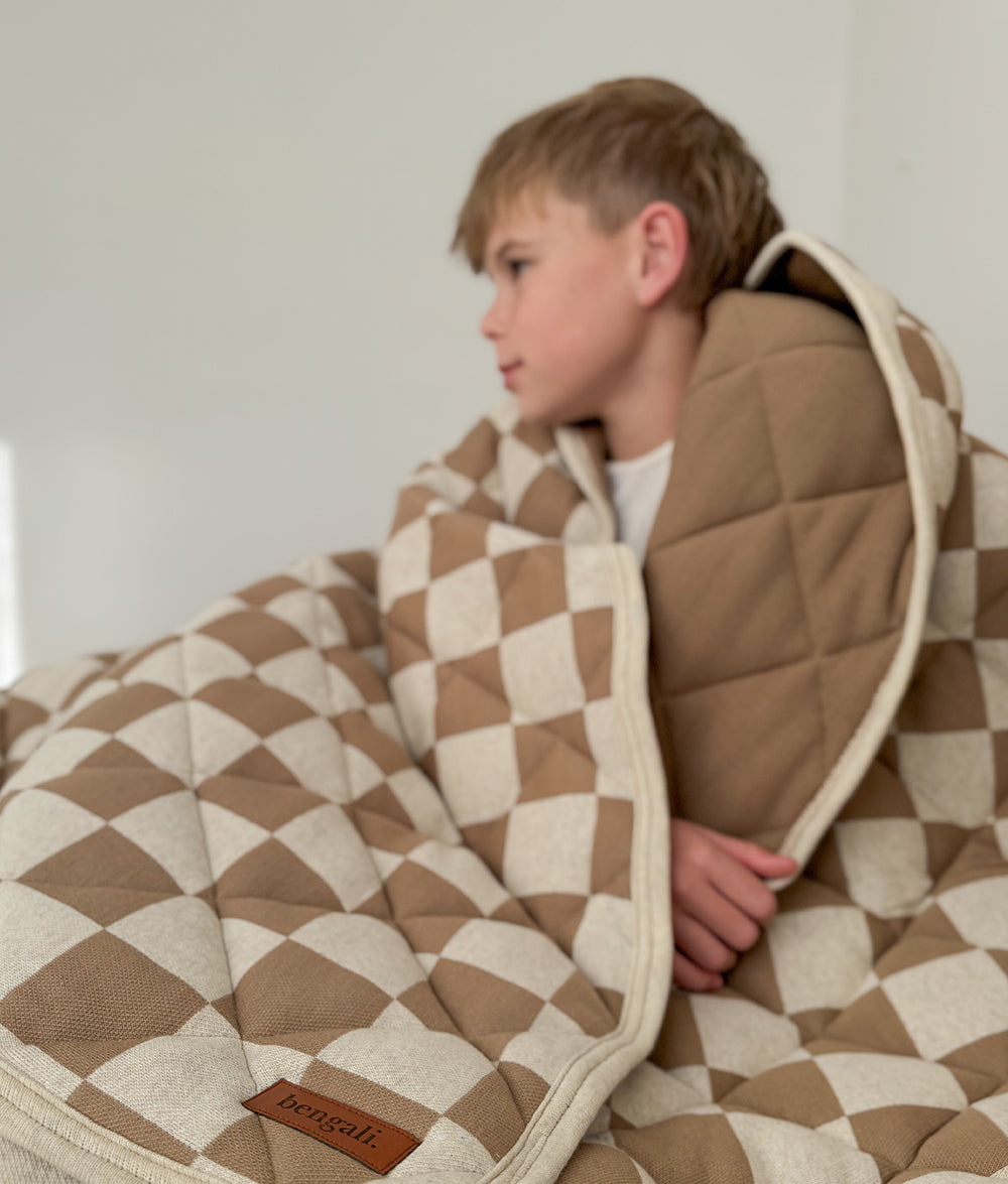 Child tucked into the Khaki Gingham reversible quilt in a warm, neutral bedroom.